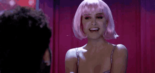 a woman in a pink wig is smiling and talking to a man in a room .