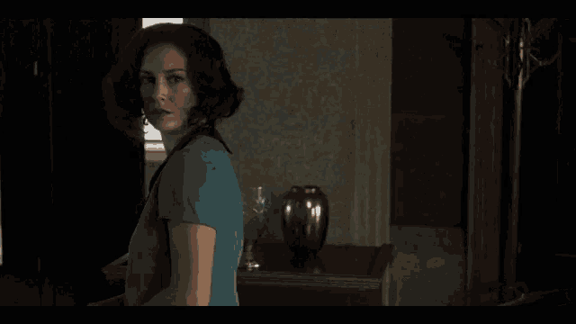 a woman in a blue shirt is standing in a room with a vase on the table .