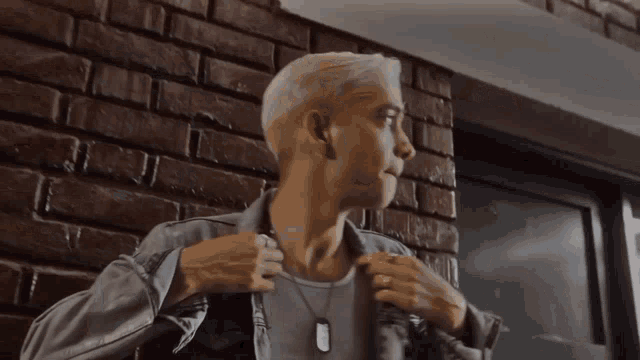 a man wearing a dog tag around his neck stands in front of a brick wall