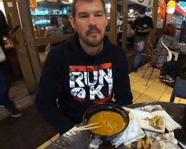 a man wearing a hoodie that says run sky sits at a table with a bowl of soup