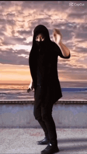 a man wearing a black hooded jacket is dancing in front of the ocean