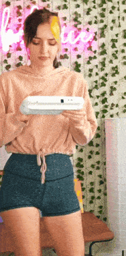a woman in a pink hoodie and blue shorts is holding a white device in her hands