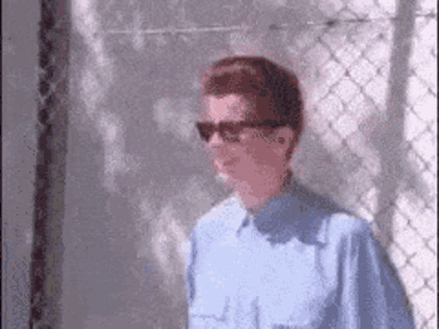 a man wearing sunglasses and a blue shirt is standing next to a chain link fence .