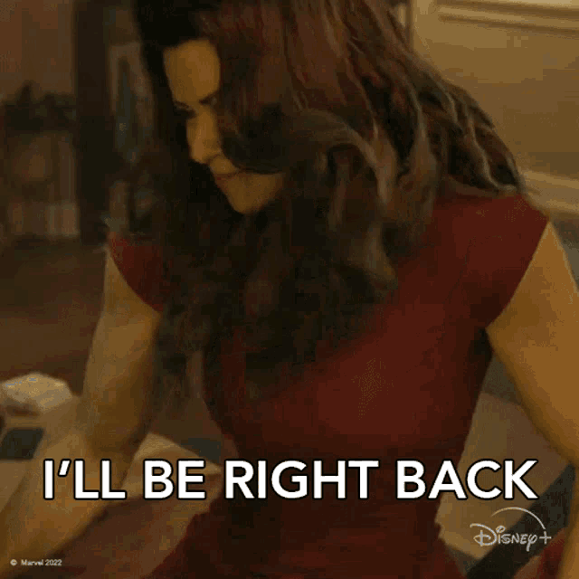 a woman in a red shirt is sitting on a bed and saying i 'll be right back