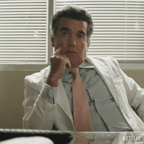 a man in a white lab coat and tie is sitting at a desk with his finger on his chin .