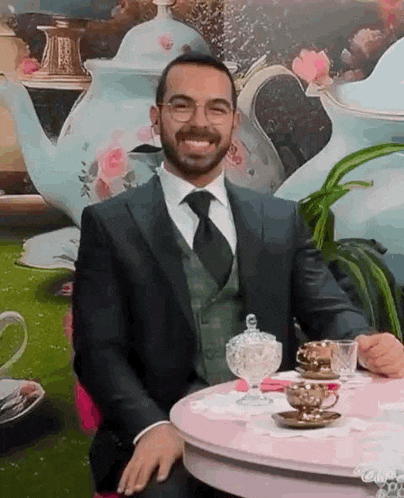 a man in a suit and tie is sitting at a table with a teapot and cups .