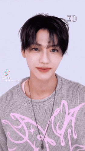 a young man is wearing a grey sweater with a pink design and a necklace