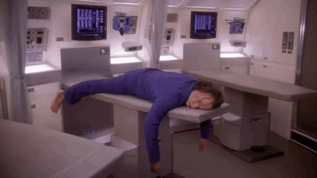 a man in a blue shirt is laying on a table with a monitor behind him that says ' emergency room '