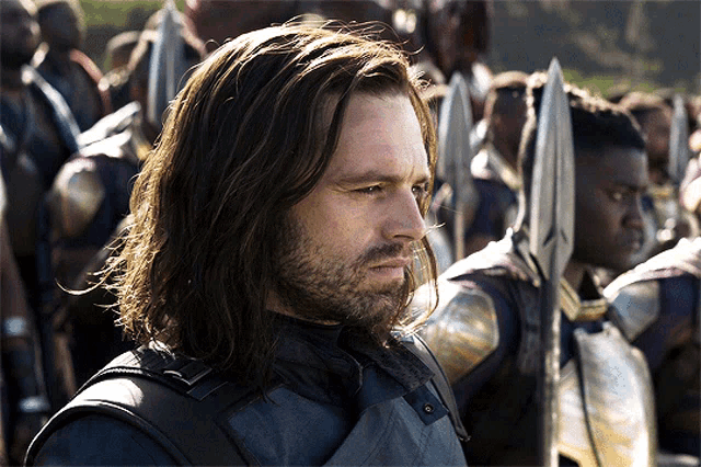 a man with long hair and a beard stands in front of a crowd of people holding spears