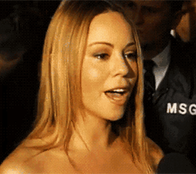 a woman is talking into a microphone with a man in a msg jacket behind her