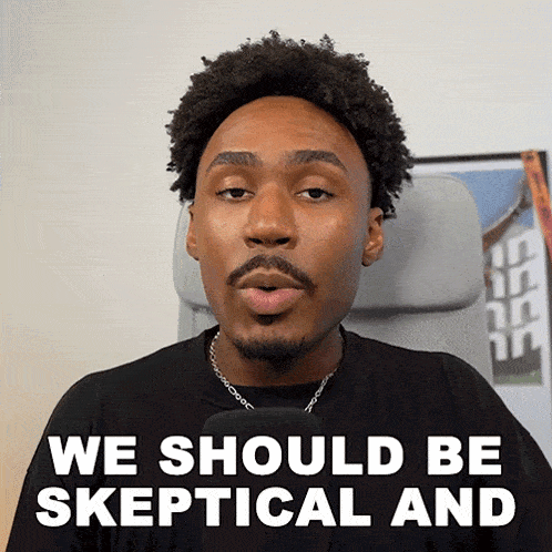 a man with a beard is talking into a microphone with the words we should be skeptical and above him