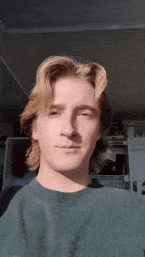a young man with long blonde hair is wearing a green sweater and looking at the camera .