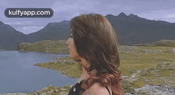 a woman is standing in front of a lake with mountains in the background and her hair blowing in the wind .