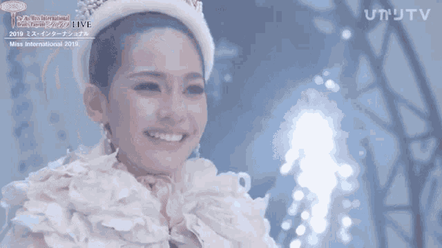 a woman wearing a crown is smiling in front of a live miss international 2019 sign