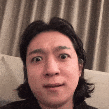a man with long hair is making a funny face while sitting on a couch in front of a curtain .