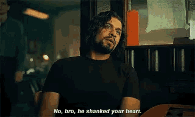 a man with long hair and a beard is sitting at a table in a room talking to another man .