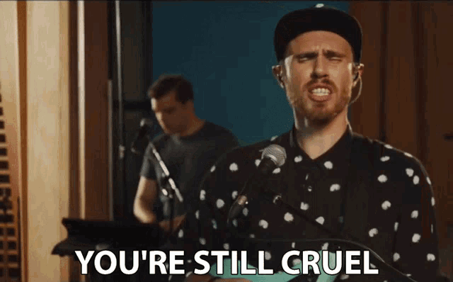 a man singing into a microphone with the words " you 're still cruel " above him