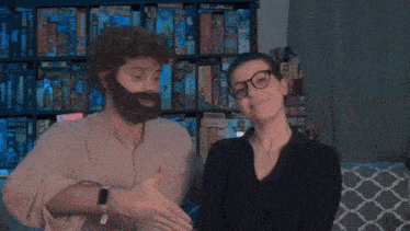 a man with a beard and glasses is talking to a woman in front of a shelf of board games