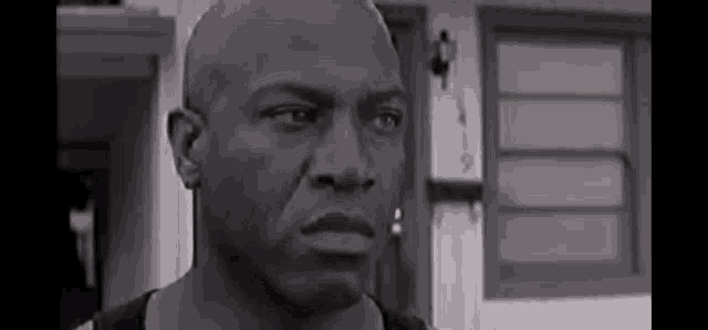 a black and white photo of a bald man standing in front of a house looking at the camera .