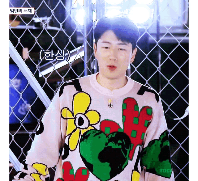 a man wearing a sweater with flowers and hearts on it is standing in front of a chain link fence