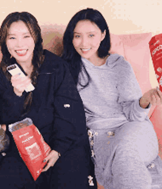 two women are sitting on a couch holding bags of popcorn and smiling