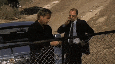 two men are standing next to a chain link fence talking on their cell phones