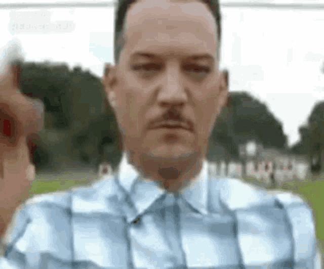 a man in a blue plaid shirt with a mustache is pointing his finger at the camera .