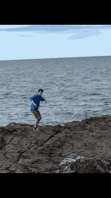 a man in a blue shirt and shorts is standing on a rocky cliff near the ocean