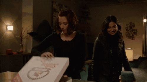 two women are standing next to each other and one of them is opening a box of sutton pizza .