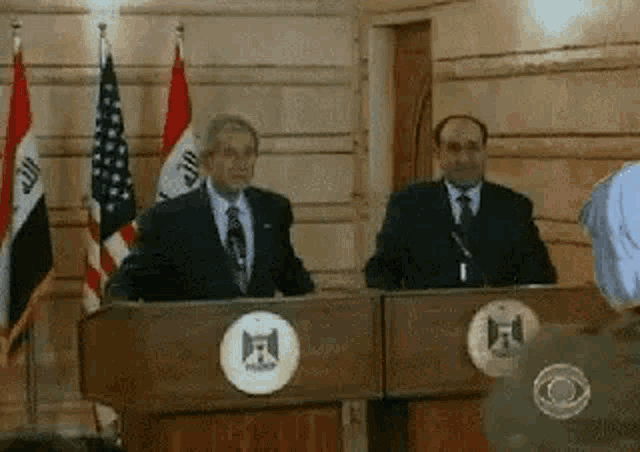 two men are standing at podiums in front of microphones in a room with flags .
