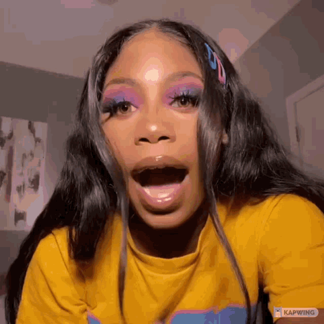 a woman wearing a yellow shirt and purple eye shadow is making a surprised face .