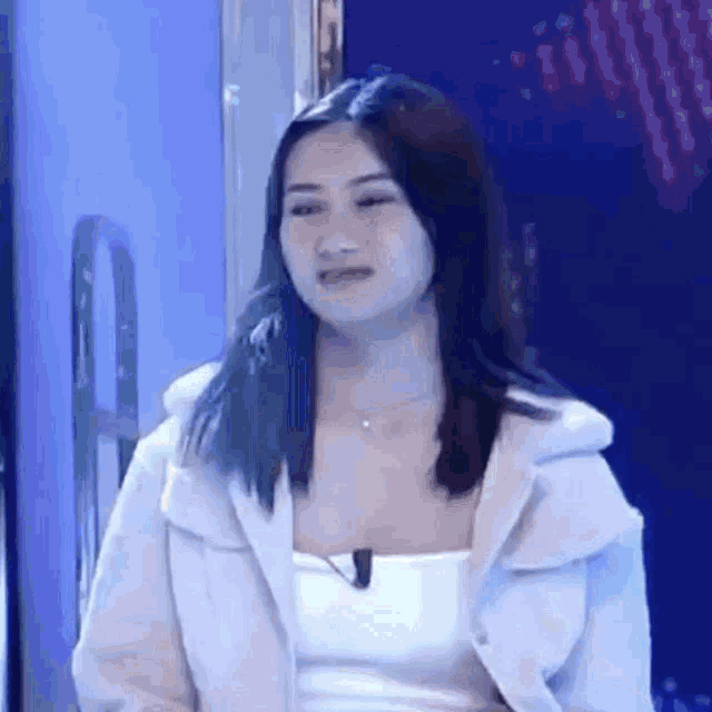 a woman in a white jacket is sitting in front of a blue wall and making a funny face .