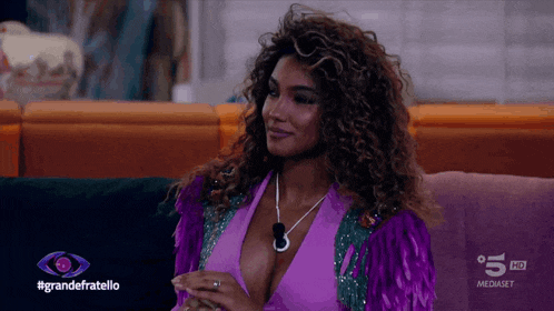 a woman in a purple top sits on a couch in front of a sign that says #grandefratello