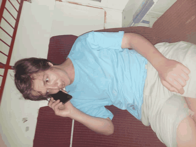 a young man in a blue shirt is laying on a bed talking on a cell phone