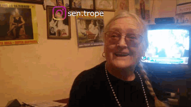 an elderly woman stands in front of a television with a sen.trope sticker on the corner