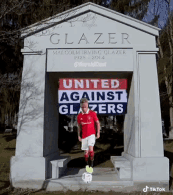 a boy stands in front of a grave of malcolm irving glazer holding a soccer ball