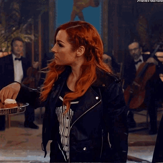 a woman with red hair is wearing a black leather jacket and holding a tray of cupcakes