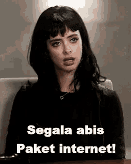 a woman with black hair is sitting at a desk with a laptop and a meme .
