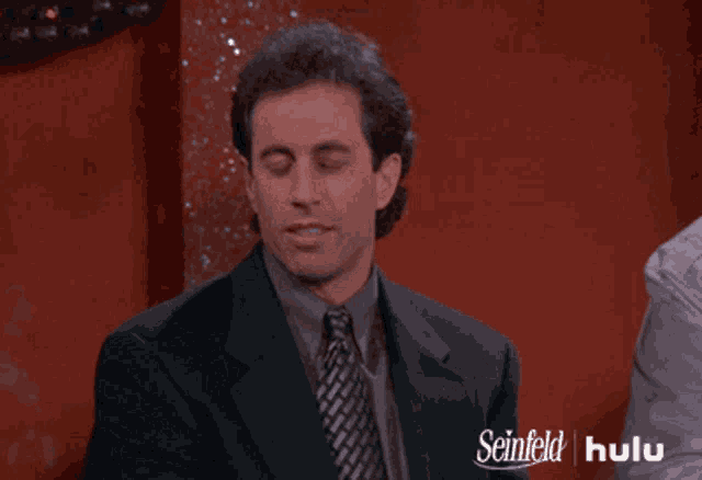 a man in a suit and tie is sitting in front of a sign that says seinfeld