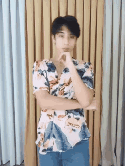 a young man in a floral shirt stands with his arms crossed and his hand on his chin