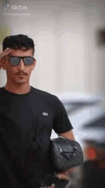 a man wearing sunglasses and a black t-shirt is holding a black bag .