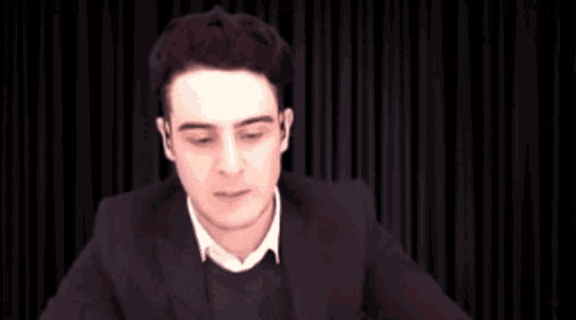 a man in a suit is making a funny face while holding a pillow in front of a black curtain .