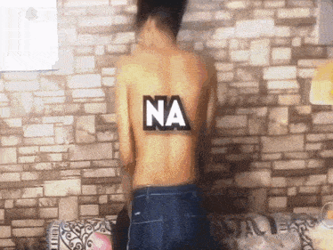 a shirtless man stands in front of a brick wall with the word na on his back