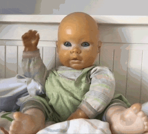 a bald baby doll with blue eyes is sitting on a bed raising its hand