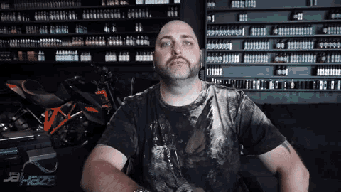 a bald man with a beard is standing in front of a shelf full of bottles of e liquid .