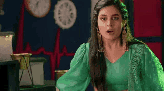 a woman in a green blouse and earrings is standing in a room with a heartbeat on the wall .