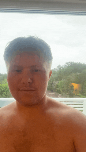 a shirtless man stands in front of a window looking out