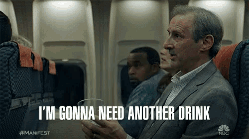 a man sitting on an airplane holding a glass and says i 'm gonna need another drink
