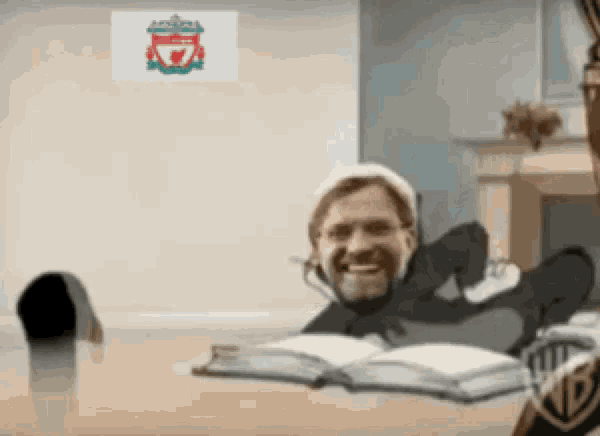 a man is sitting at a table with his feet up and a liverpool flag behind him