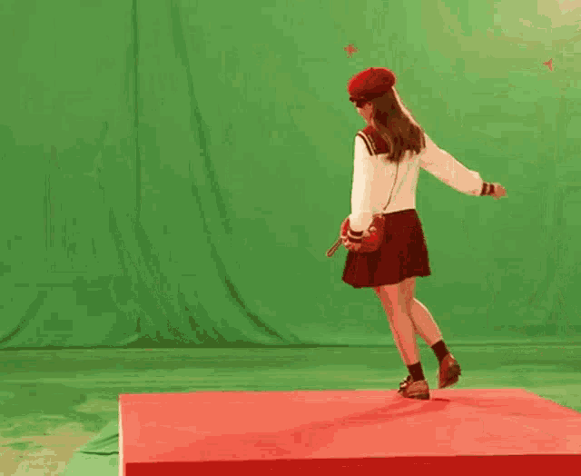a woman in a white sweater and red skirt is dancing on a red platform in front of a green screen
